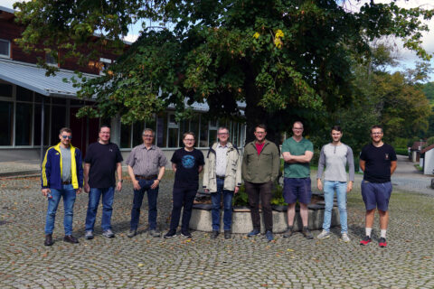 Hier ist ein Gruppenfoto zu sehen. Neun Männer stehen nebeneinander. Es sind zwei Professoren und sieben Mitarbeiter. Die Männer stehen vor einem großen Baum, und schauen locker lächelnd in die Kamera. Es ist Spätsommer.